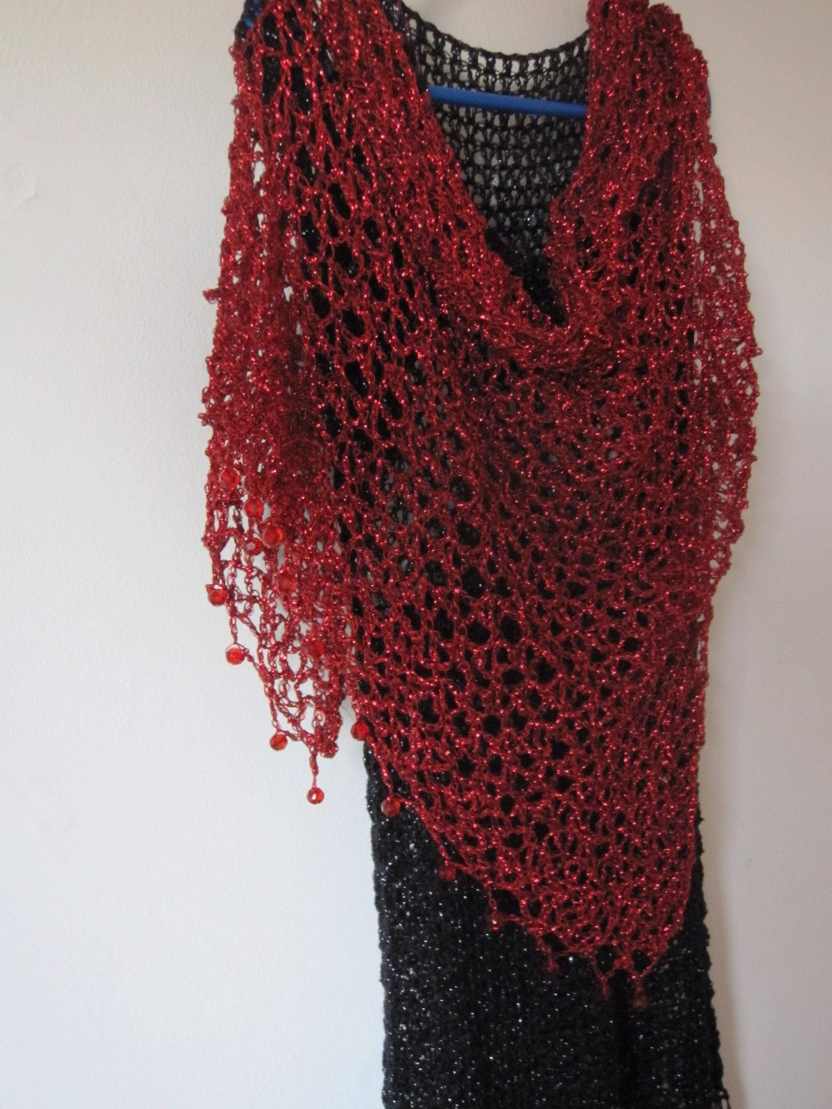 Red lurex crochet shawl draped over a black dress on a hanger, full view of the shimmering openwork wrap.